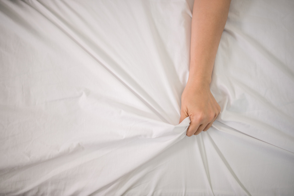 Eine Hand greift orgasmisch in die weißen Laken eines Bettes.